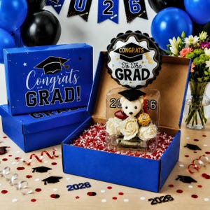 May include: A blue gift box with "Congrats Grad!" text and a graduation cap graphic. Inside, a teddy bear wearing a graduation cap, white roses, and a chocolate treat are visible. A balloon with "Congrats To The Grad" text floats above.