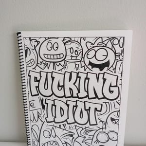 May include: A white spiral-bound notebook with a black and white cartoon illustration. The illustration features various monster-like characters and the words "FUCKING IDIOT" in bold, block letters.