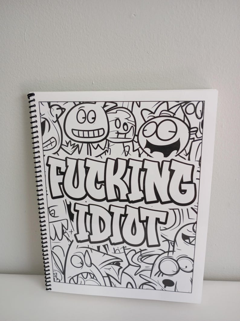 May include: A white spiral-bound notebook with a black and white cartoon illustration. The illustration features various monster-like characters and the words "FUCKING IDIOT" in bold, block letters.