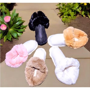 May include: A collection of plush slippers in various colors, including black, pink, tan, and white. The slippers have a soft, furry trim around the opening and a white satin footbed. The slippers are arranged on a stone surface.