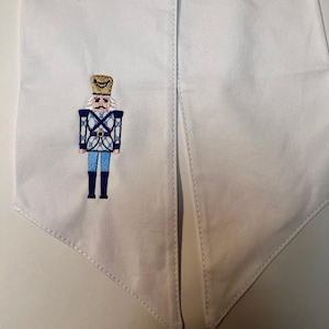 Puede incluir: Faja de tela blanca con un diseño de cascanueces bordado. El cascanueces está representado en azul, blanco y dorado, con un uniforme y sombrero detallados. La faja tiene un fondo puntiagudo, probablemente para una graduación o evento especial.