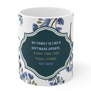 May include: A white ceramic mug featuring a dark teal scalloped frame containing the text: MY FAMILY IS LIKE A SOFTWARE UPDATE. EVERY TIME I SEE THEM, I THINK, "NOT NOW." The mug is decorated with a pattern of small blue flowers and green stems.