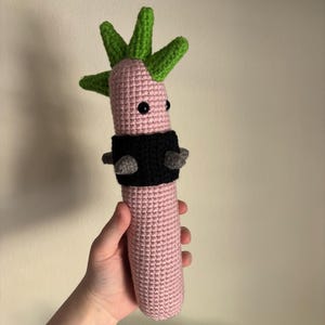 Puede incluir: Un juguete de crochet rosa con un mohawk verde y una banda negra con tachuelas plateadas alrededor de la mitad.