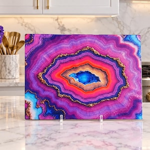 May include: A rectangular glass cutting board featuring a vibrant, multi-layered geode pattern in shades of bright pink, deep purple, and electric blue, accented with textured gold-colored metallic detailing throughout the center.