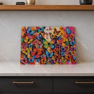 May include: A rectangular glass cutting board featuring a vibrant, multi-colored butterfly print. The design displays numerous butterflies in shades of blue, yellow, orange, and pink against a splattered, abstract paint-like background.