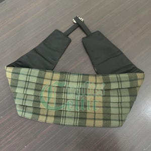 May include: A green and brown plaid kilt belt with black adjustable straps. The belt is made of a woven fabric and has a traditional plaid pattern.