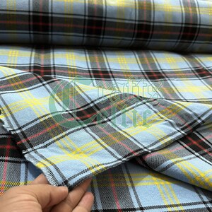 May include: Close-up of a light blue plaid fabric with black, yellow, and red lines. The fabric is folded, showing the pattern's repeating design. The fabric is likely cotton or a similar material.