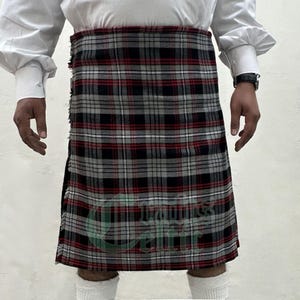 Puede incluir: Un kilt tradicional con un patrón de cuadros rojos, negros y grises. El kilt se lleva con una camisa blanca. El kilt presenta el texto "Crafts Celtic" en verde.