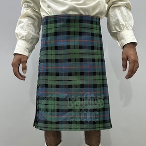 May include: A traditional kilt in a green, blue, and black plaid pattern. The kilt has a fringed edge and features the text "Crafts Celtic" in a stylized font. The garment is worn with a cream-colored shirt.
