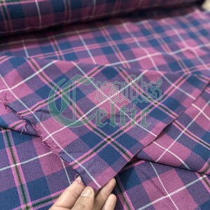 Puede incluir: Primer plano de una tela a cuadros en tonos morado, rosa y azul marino. La tela tiene un patrón de cuadros clásico con líneas de acento blancas y negras. La imagen muestra una sección doblada de la tela, revelando su textura y diseño. El texto "Crafts Celtic" es visible.