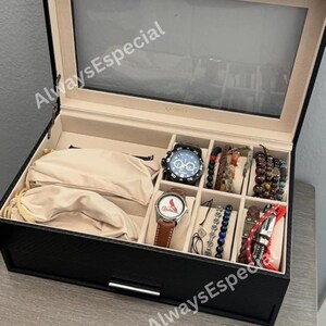 May include: Black watch box with a clear glass top, containing two watches, bracelets, and a pair of sunglasses. The watches have black and brown leather straps. The box is lined with beige fabric.