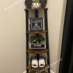 May include: A black and brown corner shelf unit with four shelves. The top shelf has two black silhouette figures and a small plant. The second shelf has a sign that reads "LADIES & GENTS". The third shelf has a sign that reads "WASH YOUR HANDS". The bottom shelf has a wire basket with two rolls of toilet paper.