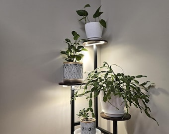 May include: A three-tiered plant stand with built-in lighting. The stand holds four potted plants, including a white ribbed pot and two pots with a black and white geometric pattern. The stand is made of dark metal and wood.