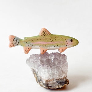 May include: A hand-painted wooden trout figurine perched on a cluster of amethyst crystals. The trout is painted in shades of green, pink, and brown with black spots.