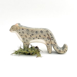 a wooden snow leopard figurine facing to the side with a long tail that curves up at the end. It is painted grey, with a white chin, chest, and belly. It has pink ears, small black details on the face and black circular spots on its body.
