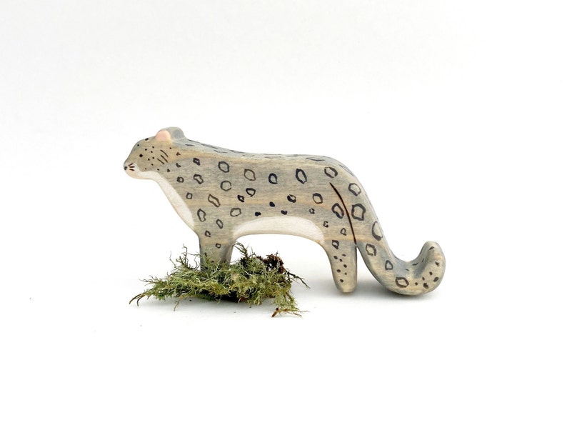 a wooden snow leopard figurine facing to the side with a long tail that curves up at the end. It is painted grey, with a white chin, chest, and belly. It has pink ears, small black details on the face and black circular spots on its body.