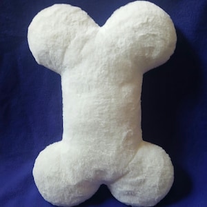 May include: A plush, white dog bone-shaped pillow. The pillow has a soft, fuzzy texture and is set against a blue background. The bone shape is a classic design, perfect for pet owners.
