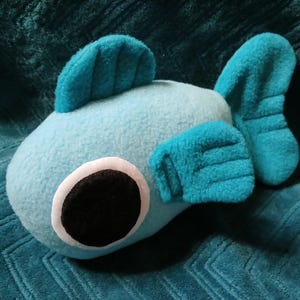 Puede incluir: Un peluche con forma de pez azul claro con aletas y detalles turquesas. El pez tiene un ojo grande y negro con un borde blanco. El juguete está hecho de tela suave y está diseñado para parecerse a un pez de dibujos animados.