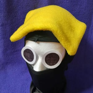 Puede incluir: Un conjunto de disfraz que presenta un sombrero de forro polar amarillo brillante, gafas grandes con montura blanca y lentes oscuras, y un pasamontañas negro. El sombrero tiene una forma puntiaguda distintiva. Las gafas son circulares.