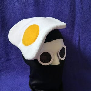 May include: A novelty costume hat resembling a fried egg, with a white beret-style top and a yellow yolk. The hat is paired with large, round, white-rimmed glasses and a black face covering, all set against a blue background.