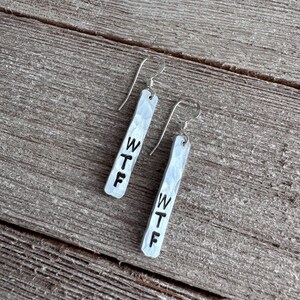 Puede incluir: Un par de pendientes de metal plateado martillado con las letras "WTF" estampadas en negro. Los pendientes cuelgan de alambres plateados y descansan sobre una superficie de madera envejecida.