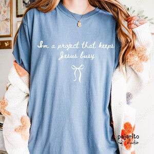 Puede incluir: Camiseta azul claro con la frase blanca "I'm a project that keeps Jesus busy" y un lazo blanco. La camiseta es de estilo casual.