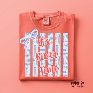 Puede incluir: Camiseta de color coral con la frase "It's River Time". El diseño presenta un fondo azul claro con rayas blancas verticales e ilustraciones de peces. Un lazo decorativo está en la parte superior izquierda.