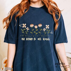 Puede incluir: Una camiseta de manga corta de color azul oscuro con un gráfico de varias flores con tallos verdes y pétalos claros. El texto "BE KIND to all kinds" está impreso en una fuente cursiva de color amarillo claro bajo el diseño floral, junto a estrellas.
