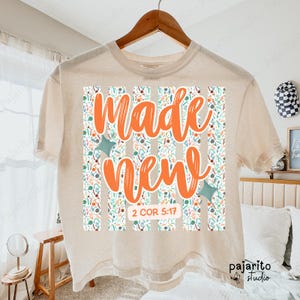 Puede incluir: Camiseta color crema con las palabras "Made New" en escritura naranja. El diseño incluye un estampado floral y el versículo bíblico 2 COR 5:17. La camiseta está colgada en una percha de madera.