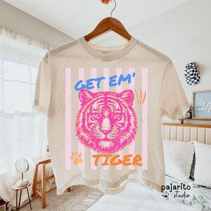 Puede incluir: Camiseta beige con un gráfico de cara de tigre rosa y las palabras "GET EM' TIGER" en azul y naranja. El diseño incluye rayas verticales rosas y blancas y una huella de pata. La camiseta está colgada de una percha de madera.