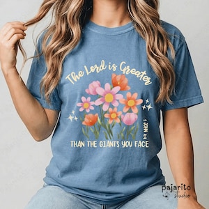 Puede incluir: Una camiseta azul de manga corta con un gráfico de flores coloridas en tonos rosa, naranja y amarillo. El texto dice The Lord is Greater than the giants you face, con la referencia 1 John 4:4 impresa verticalmente en el lado derecho.