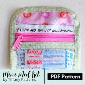 Puede incluir: Un pequeño botiquín de primeros auxilios hecho a mano con una cremallera rosa y tela estampada floral. El kit incluye vendas adhesivas de la marca Band-Aid. El texto en el kit dice "LEAP AND THE NET WILL APPEAR" y "Mini Med Kit by Tiffany Patterns PDF Pattern."