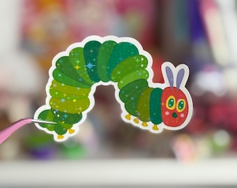 May include: A green and red cartoon caterpillar with a sparkly green body and a red head with two eyes and a smile. The caterpillar is curled into a circle.