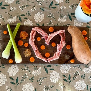 Può includere: Un tagliere in legno con manico presenta una forma a cuore fatta di carne, circondata da fette di carote e spezie. Sono presenti anche gambi di sedano e una patata dolce. Un cucchiaio decorativo e fiori completano la scena.