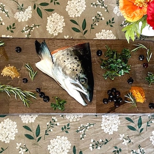 May include: A wooden cutting board with a salmon head, herbs, and spices. A decorative spoon with a house design is on the left. The scene is set on a floral patterned tablecloth. A vase of colorful flowers is in the background.