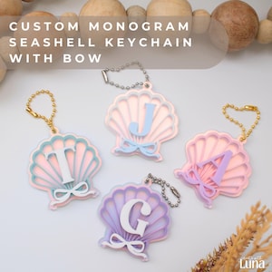 May include: Custom monogram seashell keychains with bows in pink, blue, and purple. Each keychain features a different letter, with a bow detail. The keychains have gold and silver chain attachments.