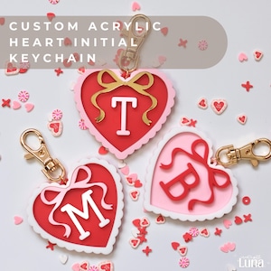 May include: Custom acrylic heart initial keychains in red, pink, and white with gold-colored clasps. Each heart features a different initial: T, M, and B. The keychains are decorated with a bow design.