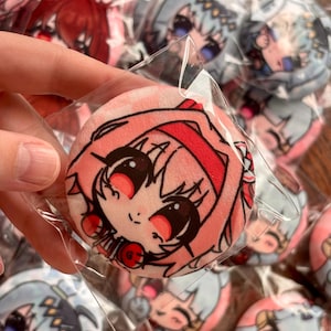 May include: A circular plush pin featuring a stylized anime-inspired character with pink hair, a red headband, and large expressive eyes. The soft fabric surface displays a vibrant graphic print of the character's face, encased in a clear plastic sleeve.