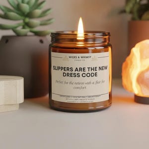 Könnte beinhalten: Eine brennende Kerze in einem bernsteinfarbenen Glas mit cremefarbenem Etikett. Das Etikett trägt die Aufschrift WICKS & WHIMSY CANDLE CO., SLIPPERS ARE THE NEW DRESS CODE, Perfect for the retiree with a flair for comfort, HANDPOURED, 100% NATURAL, NET 9 OZ.