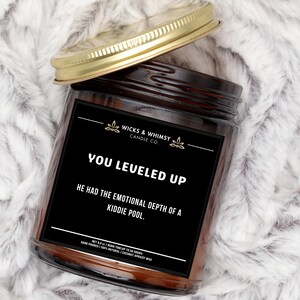 Könnte beinhalten: Ein braunes Glas-Kerzengefäß mit einem goldenen Deckel. Das Etikett ist schwarz mit weißem Text, der "Wicks & Whimsy Candle Co." und "You Leveled Up. He had the emotional depth of a kiddie pool." lautet. Das Etikett enthält auch den Text "Net 8.0 oz / Burn Time Up To 50 Hours. Hand Poured 100% Natural | Coconut Apricot Wax."