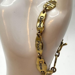 Pode incluir: Pulseira dourada com elos ovais inscritos com palavras como "Believe", "Giving" e "Dream". A pulseira é exibida em uma mão de manequim branca, mostrando seu design e mensagem. A pulseira tem um fecho.
