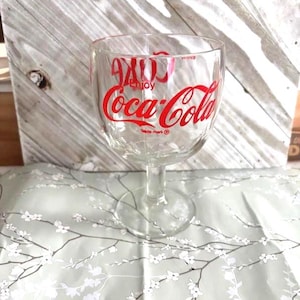 Pode incluir: Um copo de vinho Coca-Cola transparente com o logotipo da Coca-Cola em vermelho. O copo tem uma taça larga e uma haste fina. As palavras "Enjoy Coca-Cola" estão impressas no vidro. O copo está sobre uma superfície com padrão floral.