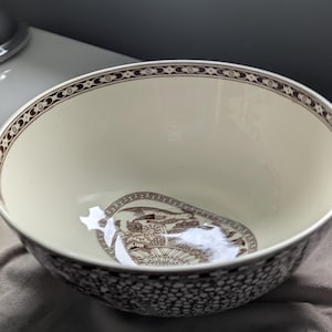 May include: A cream-coloured ceramic bowl featuring a dark brown decorative border along the rim and a detailed brown illustration of a bird perched on a floral basket at the bottom. The exterior of the bowl displays a dense, speckled pattern.