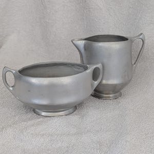 May include: A vintage silver-colored creamer and sugar bowl set. The creamer has a handle and a spout, while the sugar bowl has two handles. Both pieces have a simple, classic design and sit on a white surface.