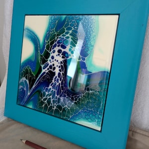May include: Square abstract art piece in a teal frame. The artwork has a fluid design with shades of blue, green, and white on a white background. A brown pencil is in front of the artwork. The frame is painted teal.