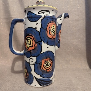May include: A tall ceramic coffee pot with a lid, handle, and spout. The pot is white with blue floral designs and orange centres. The handle and spout are blue. The lid has a yellow rim.