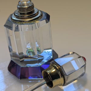 Può includere: Un flacone di profumo in vetro trasparente sfaccettato, di forma esagonale con base viola intenso. Il flacone presenta un colletto a vite in metallo dorato. Un tappo in vetro abbinato con chiusura in metallo dorato e bacchetta applicatrice è accanto.