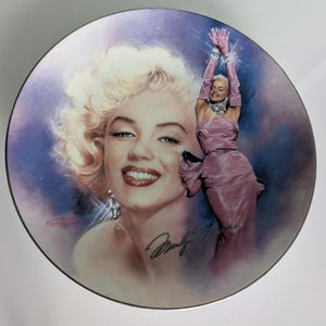 Puede incluir: Un plato decorativo con un retrato de Marilyn Monroe con su firma. El plato tiene una segunda imagen en relieve de Marilyn con un vestido y guantes rosas. El fondo es un degradado de morado y azul.