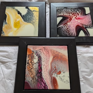 May include: Three framed abstract art pieces. The art features fluid acrylic pours in shades of yellow, red, pink, and white. The frames are black, and the art is displayed on a white surface. Each piece is square, with a dark, matte black frame.