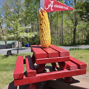 Könnte beinhalten: Ein roter, hölzerner Miniatur-Picknicktisch mit einem Maiskolben darauf. Der Tisch steht auf einer Holzoberfläche. Im Hintergrund ist eine rot-weiße Flagge mit dem Buchstaben "A" und den Worten "Crimson Tide" zu sehen.